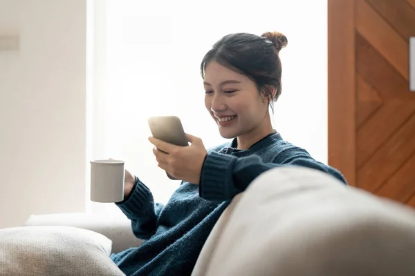 Alegre jovem mulher asiática usando telefone celular enquanto sentado em um sofá em casa. — Fotografia de Stock