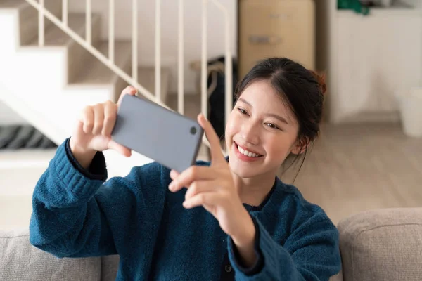Foto van jonge aziatische vrouw het nemen van selfie op mobiele telefoon terwijl zitten op de bank in helder appartement — Stockfoto