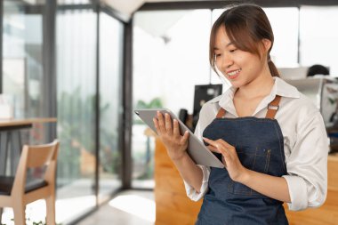 Kahve dükkanının arka planındaki müşteriden gelen siparişleri kontrol etmek için dijital tablet tutan Asyalı kadın barista, KME iş konsepti