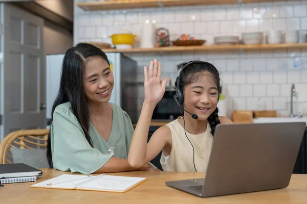 Happy asian mother and child sitting at kitchen table with colored pencils, attending virtual drawing class via video call, smiling and waving hello at laptop computer screen to greet online teacher