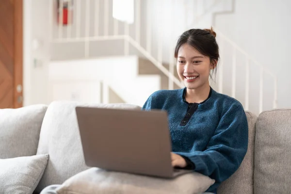 Glücklich casual beautiful young asian woman working on a laptop sitting on the couch im the house. — Stockfoto