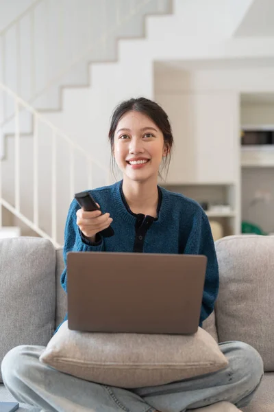 Linda mulher asiática sentada no sofá na sala de estar usando laptop e assistindo algo interessante na televisão — Fotografia de Stock
