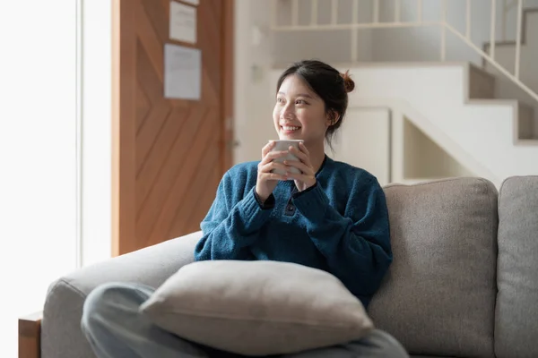 Feliz jovem mulher asiática sentada em um sofá com uma xícara de café em casa — Fotografia de Stock
