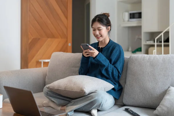 Attraktive lächelnde Frau mit Smartphone, während sie zu Hause auf dem Sofa sitzt. Kommunikations- und Gemütlichkeitskonzept — Stockfoto
