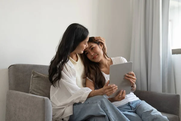 Feliz pareja lesbiana abrazándose a divertirse usando tableta digital relajándose en el sofá en casa. Dos sonrientes mujeres asiáticas amigas sosteniendo la computadora mirando la pantalla disfrutando de navegar online viendo videos. —  Fotos de Stock