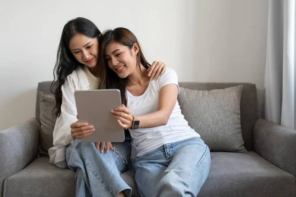 Feliz joven pareja de lesbianas abrazándose a divertirse usando tableta digital relajándose en el sofá en casa. Dos sonrientes mujeres asiáticas amigas sosteniendo la computadora mirando la pantalla disfrutando de navegar online viendo videos —  Fotos de Stock