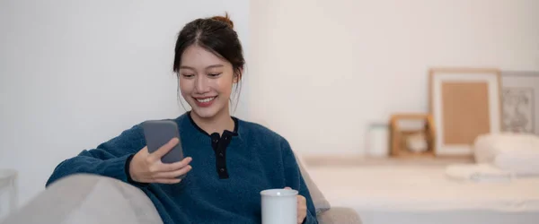 Счастливая молодая азиатка, сидя дома на диване, пользуется мобильным телефоном — стоковое фото
