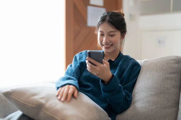 Happy casual beautiful asian woman is talking on ein phone sitting on a sofa at home. — Stockfoto