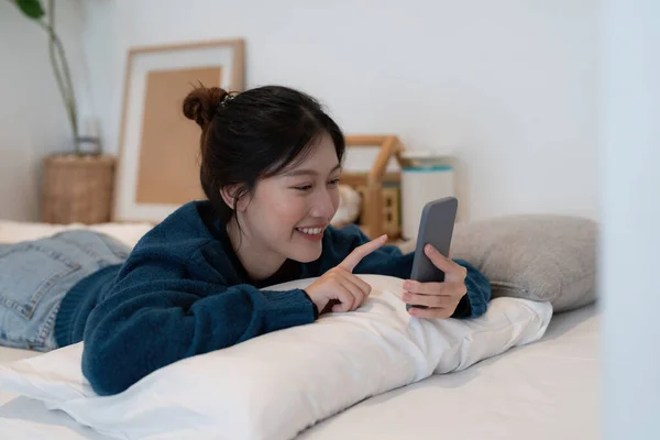 Schöne asiatische Frau legt sich mit Handy auf weißes Bett und lächelt am Sonntag in der Früh. — Stockfoto