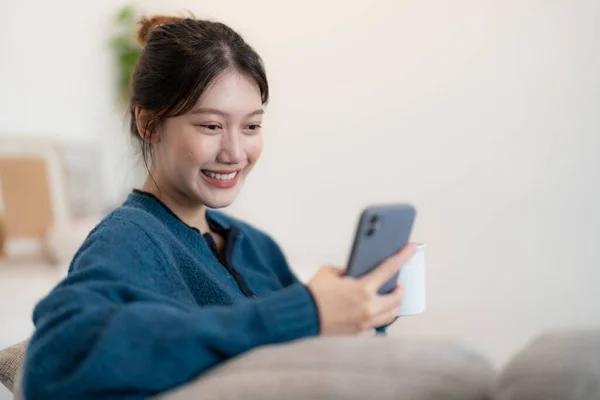 Gelukkig jong aziatisch vrouw met behulp van mobiele telefoon terwijl zitten een bank thuis — Stockfoto