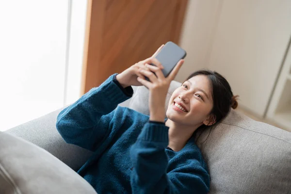 自宅のソファに横になりながら、携帯電話を使って魅力的なアジアの女性を笑顔のイメージ — ストック写真