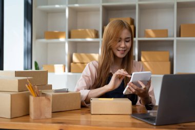 Genç bayan küçük işletme sahibi evden online alışveriş yapıyor. Arka planda paket ürün hazırlayan cep telefonlu müşterilerin siparişlerini doğrulayın. KOBİ girişimcisi veya serbest yaşam tarzı kavramı