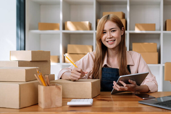 Portrait young attractive asia female owner startup business look at camera work happy with carton box at home prepare delivery in sme supply chain, procurement, omnichannel ecommerce online concept
