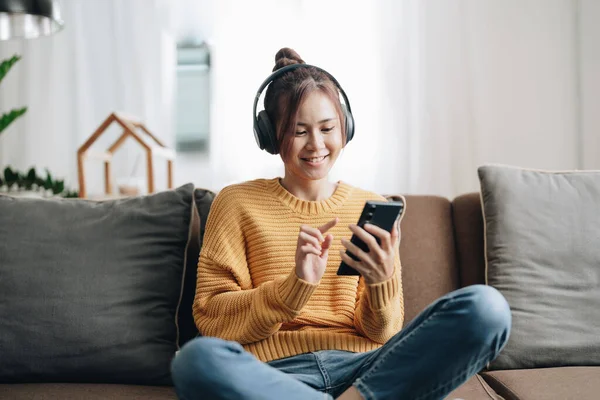 stock image Freelance asian woman with mobile phone listening music in headphones and relax at home. Happy girl sitting on couch in living room