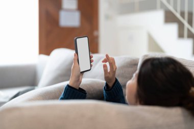 Mutlu genç Asyalı kadın oturma odasında kanepede uzanıyor ve evde cep telefonu kullanıyor..