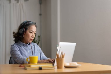 Olgun Asyalı kadın kulaklık takıyor, dizüstü bilgisayar kullanıyor, video görüşmesi yapıyor, mutfakta masada oturuyor, gözlüklü hoca çevrimiçi konferans veriyor, Webinar kaydediyor, öğretmenlik yapıyor..