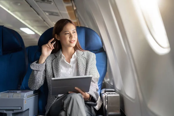 Successful beautiful young asian business woman sits in airplane cabinplane and works on digital tablet with stylus. Flying at first class