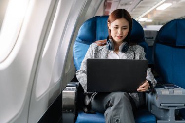 Kulaklık takan Asyalı genç bir kadın uçuş, seyahat ve iş konsepti sırasında birinci sınıfta pencerelerin yanında oturuyor..