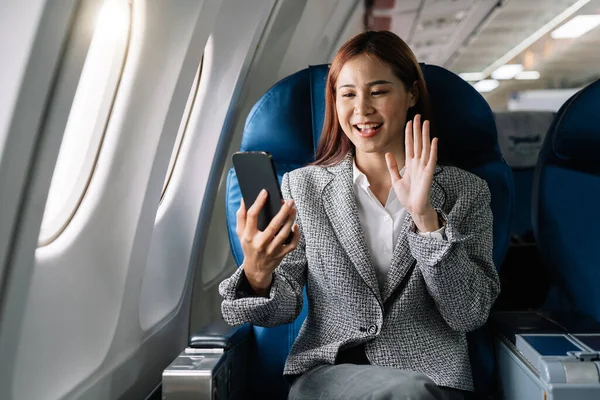 Smiling young asian businesswoman waving hand during video call on smartphone in plane.