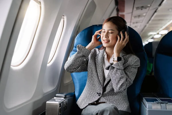 Young asian woman in wireless headphones listening to music and smiling during fly in airplane, travel concept.