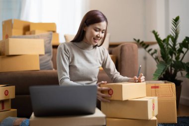 Asyalı kadın çevrimiçi mağaza küçük işletme sahibi girişimci kargo ecommerce kutusunu paketliyor web sitesi perakende siparişini kontrol ediyor masadaki dizüstü bilgisayar hazırlık paketini kullanarak. İndirme konsepti