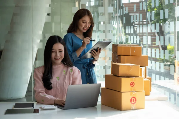 Young women teamwork partners packing order into box together using touchpad to do delivery to client. two asian female colleagues with tablet checking goods number and prepared cardboards in office