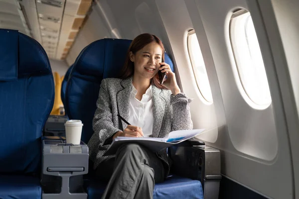 Young attractive and successful business asian woman talking on the phone and working while sitting in the chair of his private business plane.