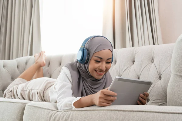 full length view of young muslim woman laying on couch in living room and watching movie on digital tablet.