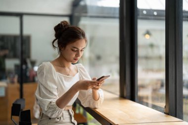 Kafede akıllı telefon kullanan Asyalı genç bir kadın. Evde akıllı telefon kullanan, mesajlaşan ya da sosyal ağları karıştıran gülümseyen bir kadın