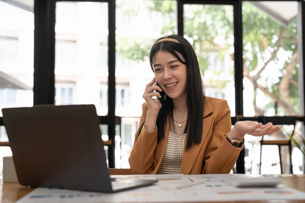 Neşeli Asyalı kadın çalışan cep telefonuyla konuşan bilgisayarla çalışıyor. Memnun iş kadını şirket müşterisiyle telefonda mali rapor ya da proje detaylarını tartışıyor.