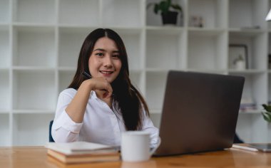 Ofiste otururken gülümseyen ve dizüstü bilgisayarla çalışan Asyalı neşeli kadın işçinin görüntüsü..