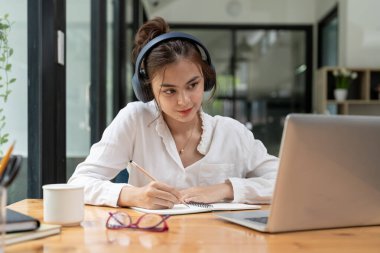 Online education, e-learning. young woman studying remotely, using a laptop, listening to online lecture, taking notes while sitting at home