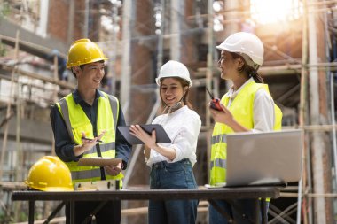 architector and engineers with draft plan of building and tablet computer talking on constructing site