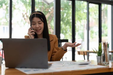 young asian woman talking on the mobile phone and smiling while sitting at her working place in office