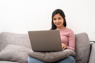 cheerful asian woman using laptop while sitting on sofa in living room.
