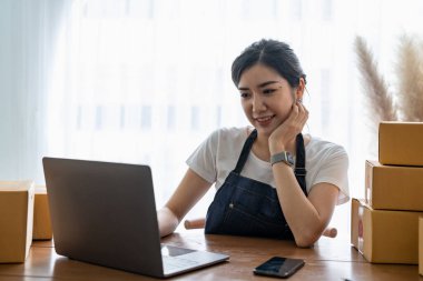Genç bayan küçük işletme sahibi evden online alışveriş yapıyor. Arka planda paket ürün hazırlayan cep telefonlu müşterilerin siparişlerini doğrulayın. KOBİ girişimcisi veya serbest yaşam tarzı kavramı