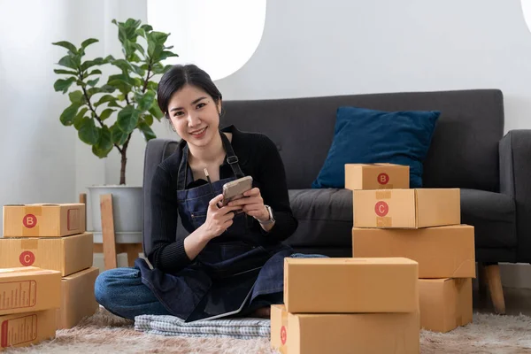 Portrait business Asian woman using mobile phone checking information on parcel shipping box before send to customer. Entrepreneur small business working at home