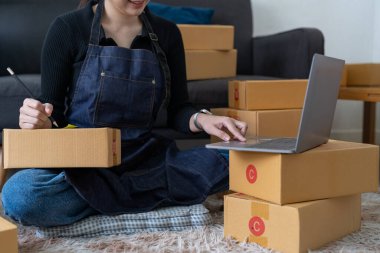 startup small business owner writing address on cardboard box at workplace. Freelance Asian woman small business entrepreneur SME working with box at home