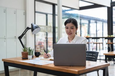 Asian woman writing and taking note on notebook with laptop in office