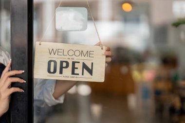 Small business owner smiling while turning the sign for the opening of the place after the quarantine due to covid-19. Close up of woman hands holding sign now we are open support local business