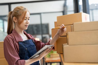 Genç, çekici Asyalı kadın şirket sahibi kameraya bakın. Evde kutuyla mutlu bir şekilde çalışıyor. Malzeme tedarik zincirinde paket teslimatı hazırlıyor..