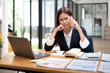 Young serious office worker female sitting at desk at workplace holding mobile phone makes business call listens client claims feels displeased annoyed and anxious, problems at work concept.