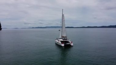 Phuket, Tayland, 05 Eylül 2020: Açık yelkenli beyaz yelkenli katamaran, tropikal bir adanın yanında duruyor, geminin etrafında insansız hava aracı var.