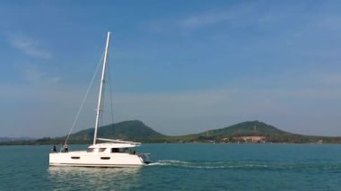 Yelkensiz bir katamaran denizde hareket eder, bir yelkenli ada boyunca deniz boyunca gider.