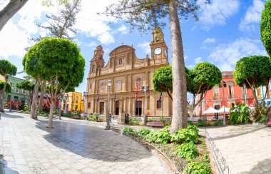February 20 2022-Galdar, Spain : Historical center in sunny weather, with old church and green palm trees , beautiful fountain in front clipart