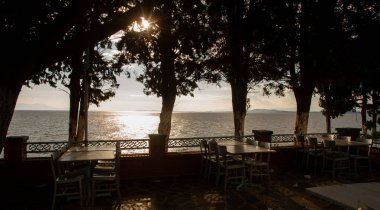 Bir Yunan tavernasının terasından gün batımının resmi, arka planda denizi ve Yunan adalarını görebilirsiniz ve ön planda güneş ışınlarının geçtiği sandalye ve ağaçları görebilirsiniz..
