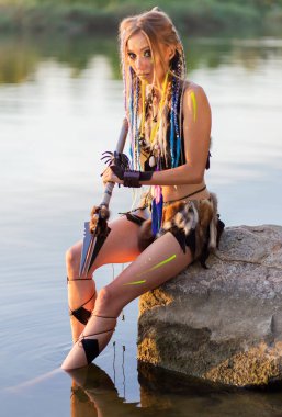 Ortaçağ kürklü bir kadın elinde mızrakla, savaş boyasıyla taş bir taşa yaslanmış..