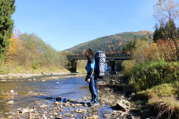 a tourist with dreadlocks and a hiking backpack on his back crosses a mountain river into a ford, against the backdrop of the Carpathian mountains