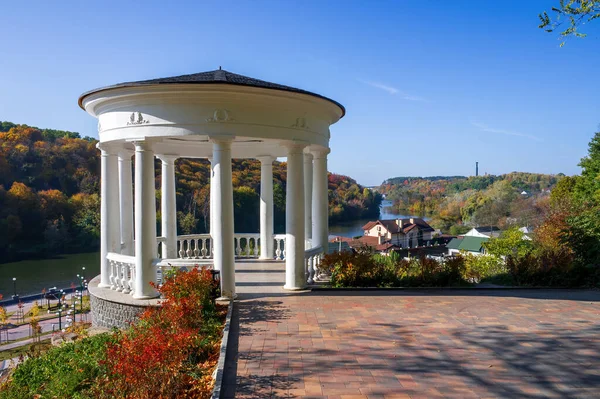White gazebo in city park of Zhytomyr, Ukraine at sunny autumn day. Picturesque view on colourful hillside on the banks of Teteriv river