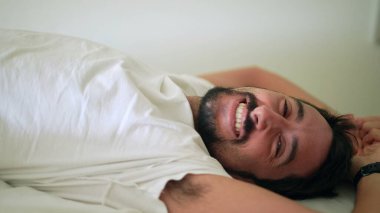 Genç adam yatakta uzanmış kameraya bakıp gülümsüyor.